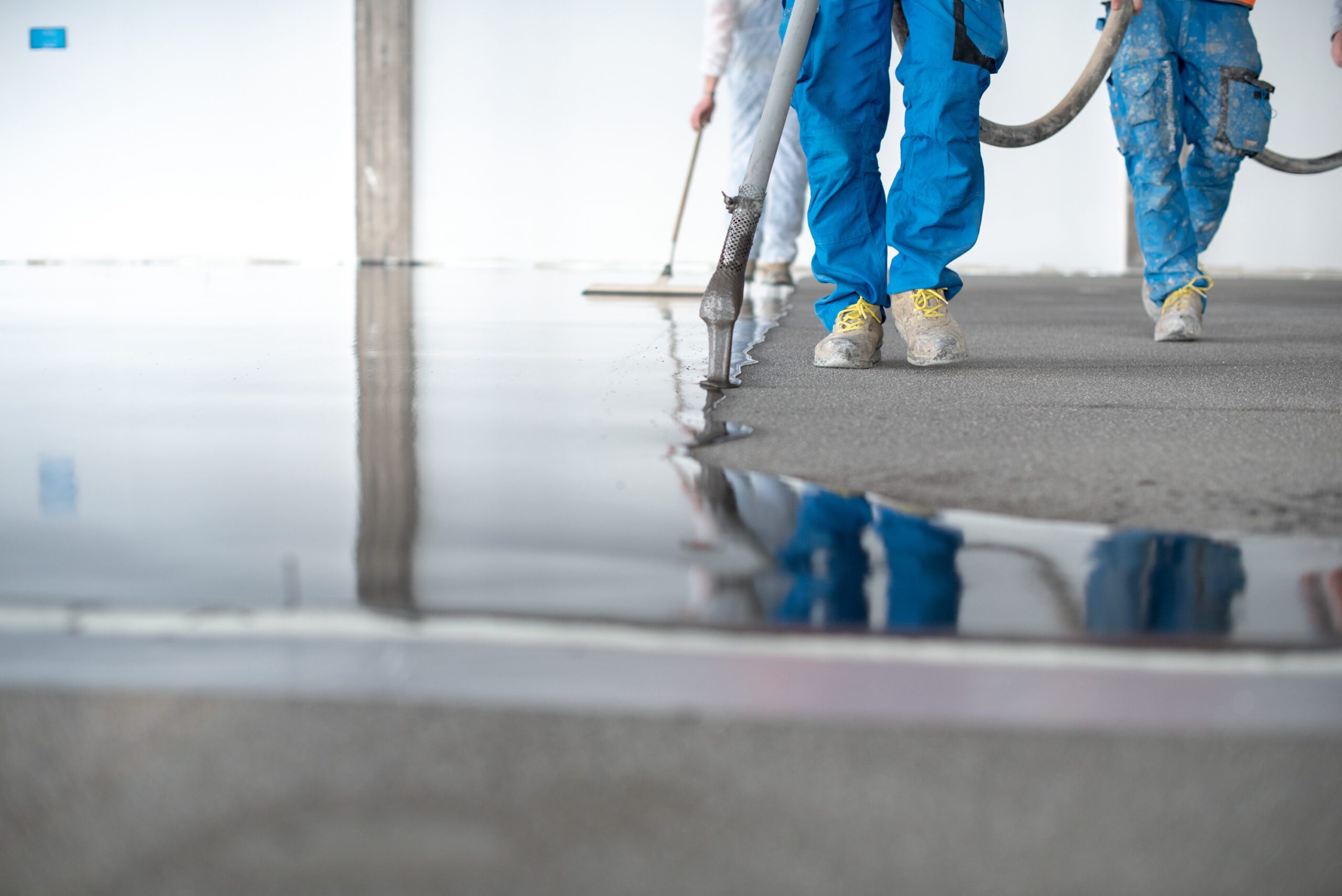 Ground level of faceless site constructors in overall holding hose while pouring liquid self leveling concrete on floor in building