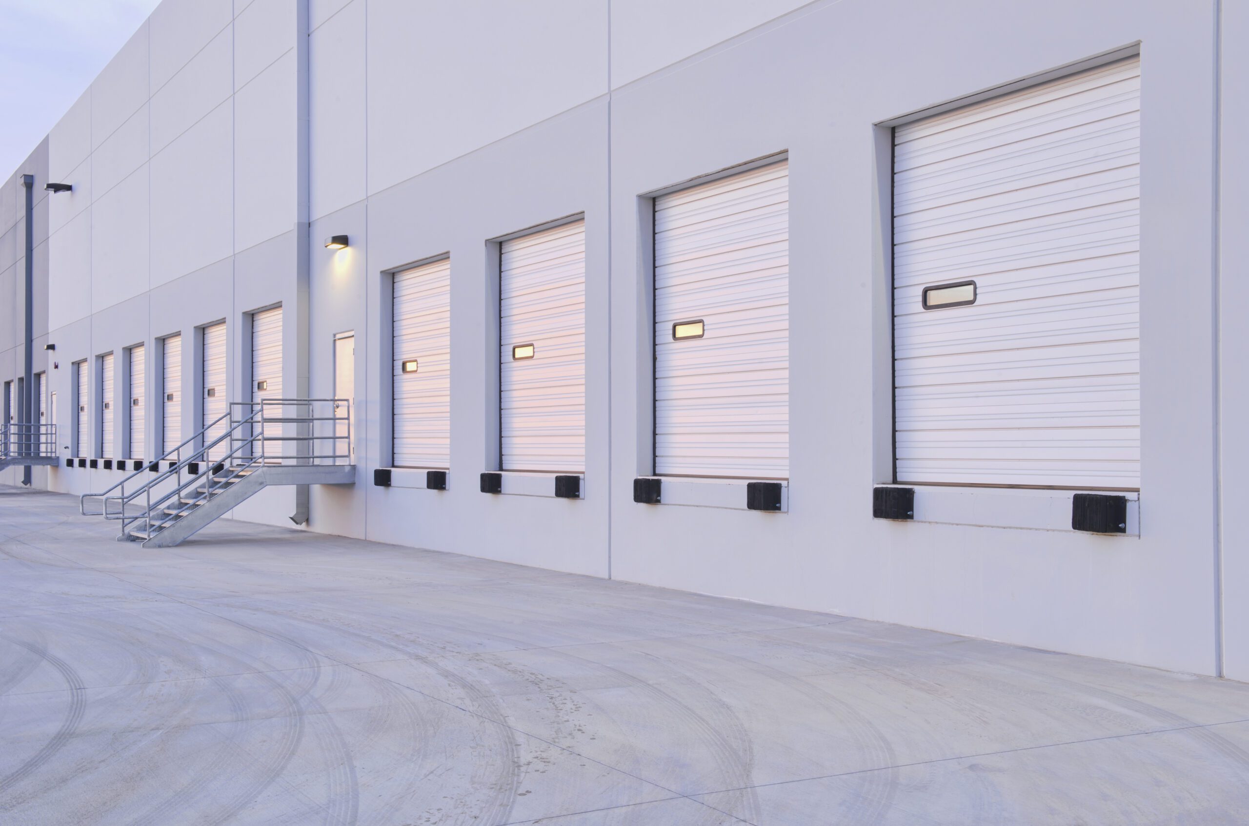 Exterior of a modern distribution center with multiple closed white roll-up bay doors and concrete loading area.