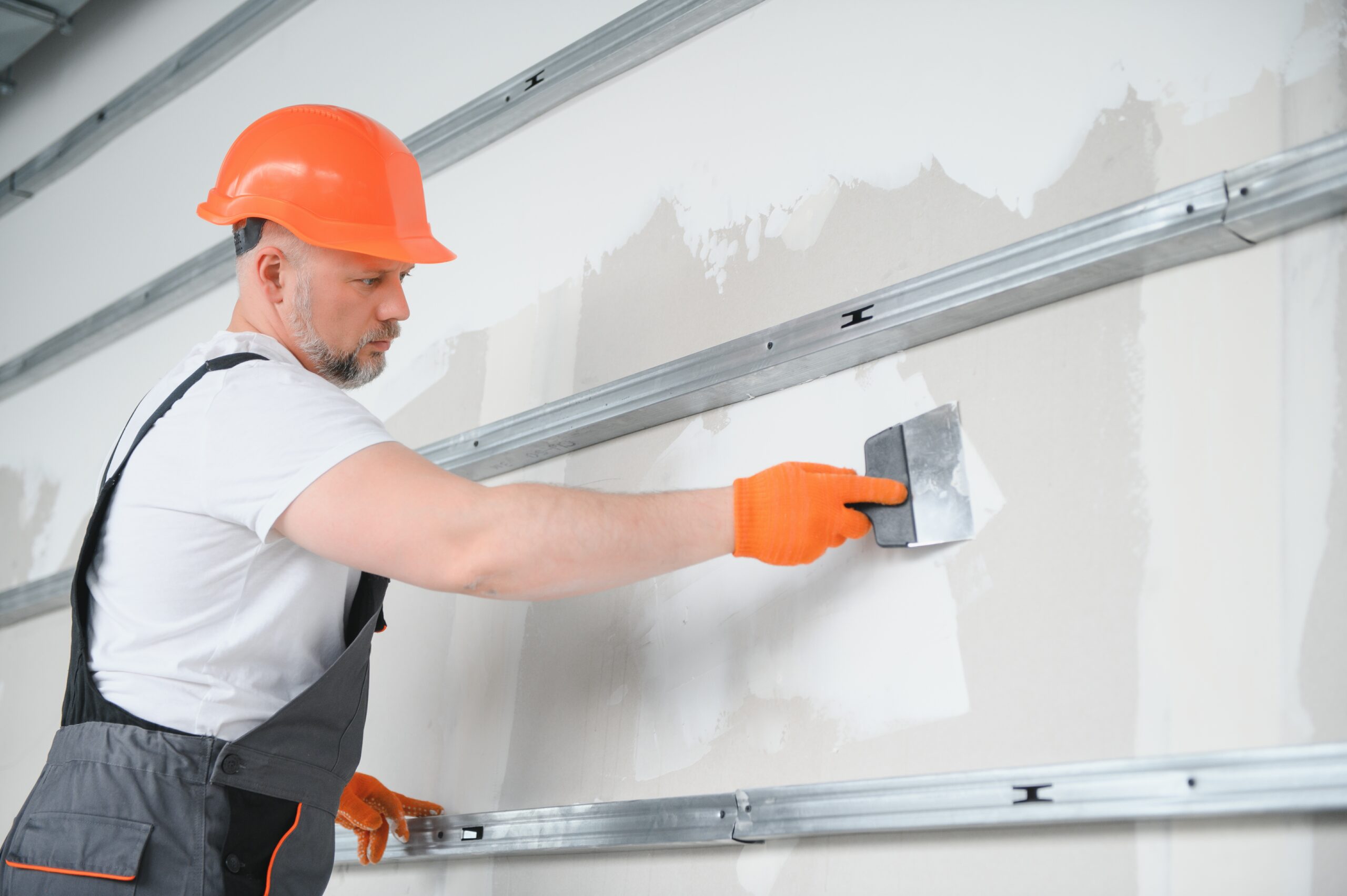 Male construction worker in hard hat using a putty knife to smooth drywall joint compound between panels.