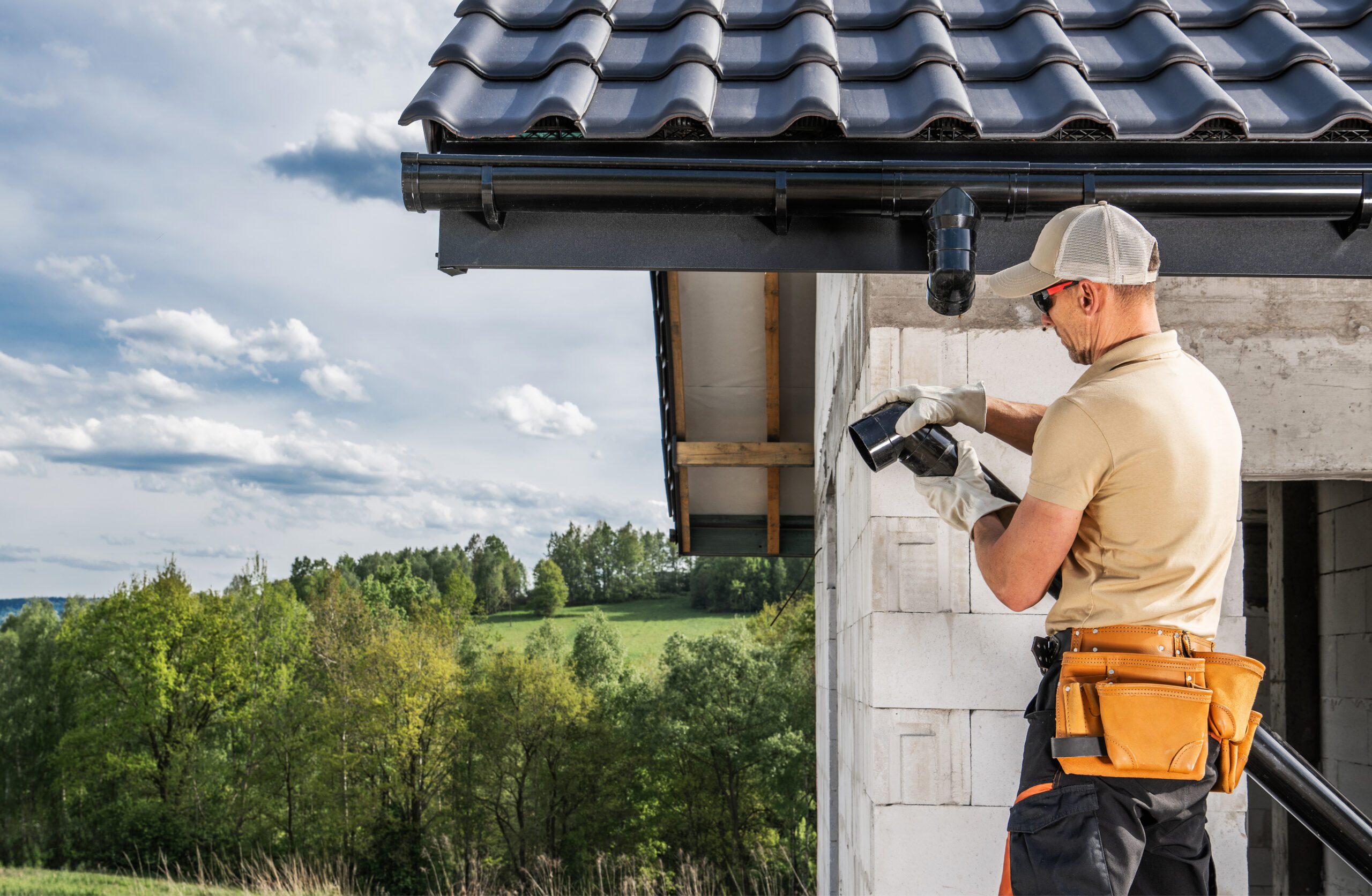 Worker installing rain gutters on new house construction with trees in background