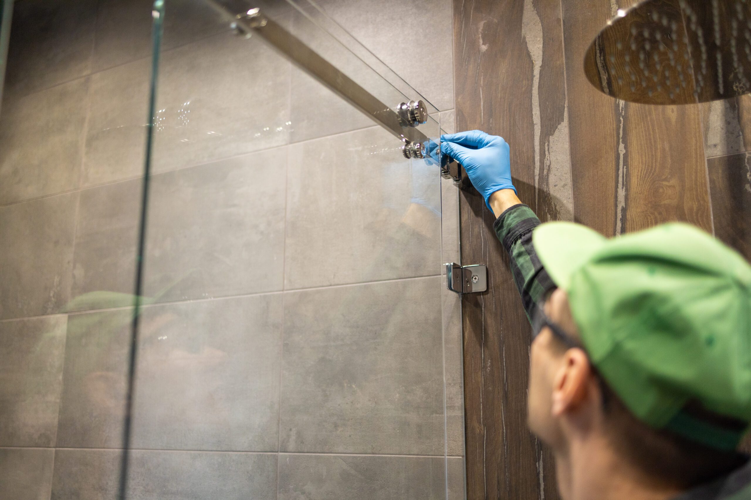 Technician adjusting fittings on a frameless glass shower door with wood-look tile background