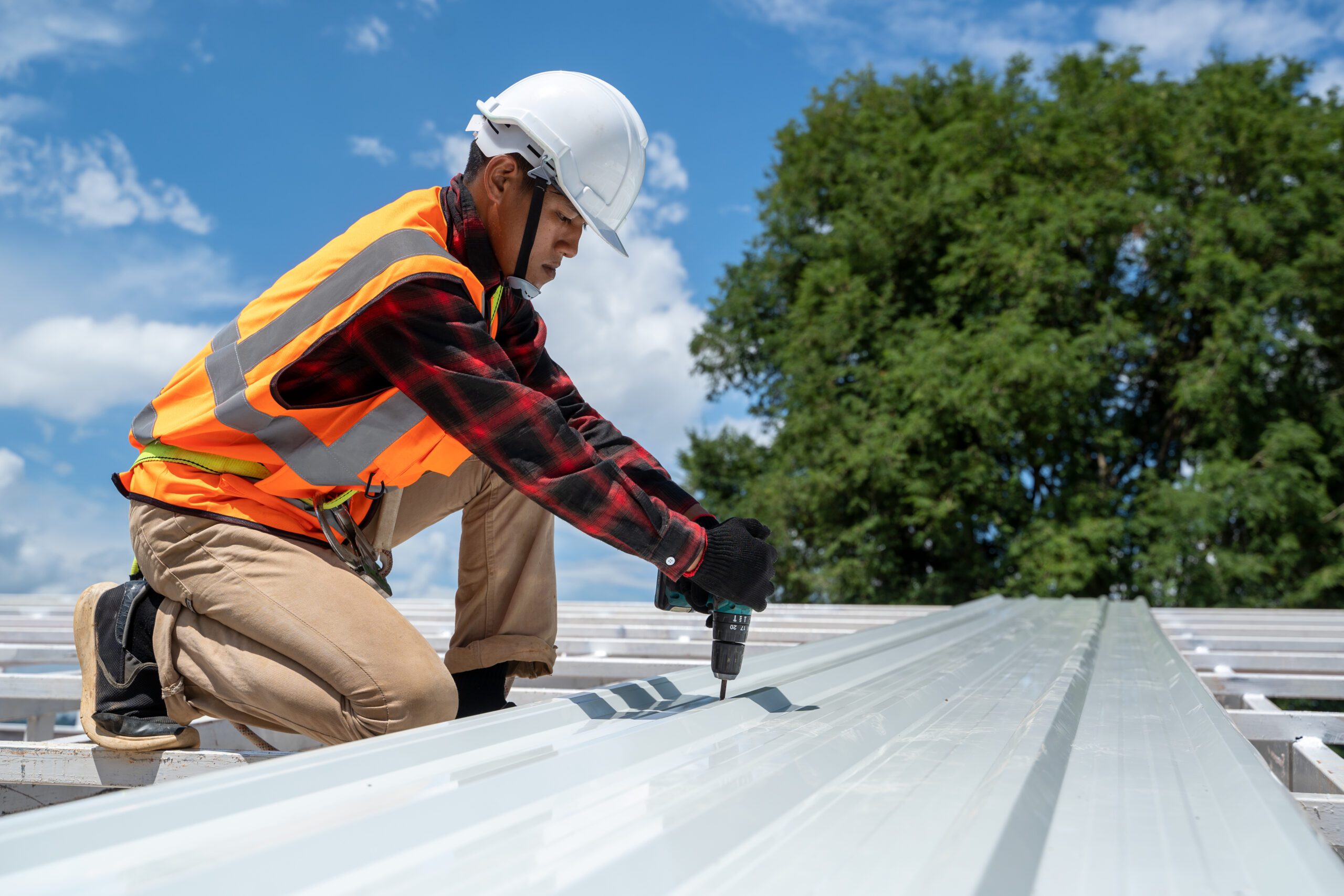 Roofer in safety gear drilling into white metal roofing during installation on a bright sunny day